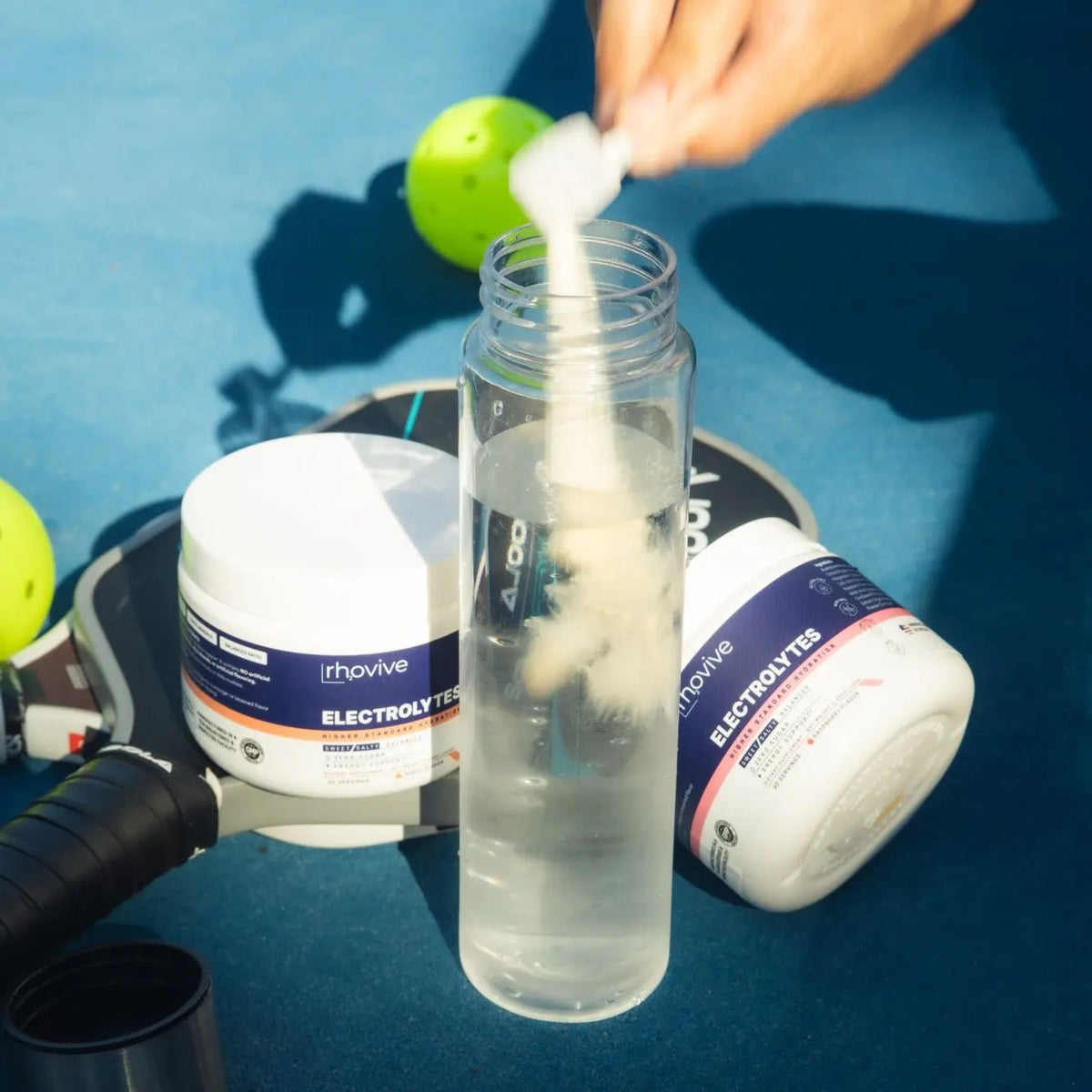 A scoop of Rhovive poured into a water bottle next to a jar of Peach and a jar of Raspberry and a pickleball paddle