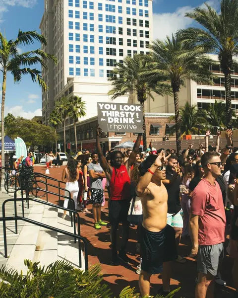 A crowd at an outdoor fitness event; a sign reads: You look thirsty! Drink Rhovive