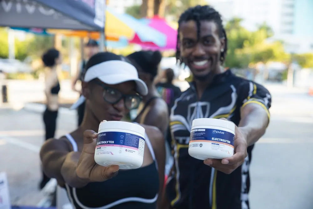 Two people at an outdoor event, each holding a jar of Rhovive toward the camera