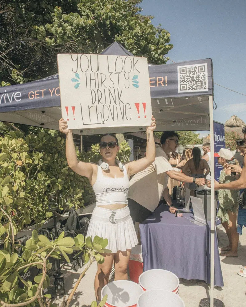 At the Rhovive tent, a woman holds up a sign: You look thirsty, drink Rhovive