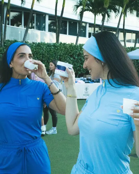 Two women in matching blue sets sipping Rhovive samples at an event